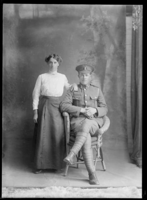 Studio portrait of an unidentified man and woman, showing the man, sitting, dressed in military uniform, and the woman, dressed in long skirt and blouse, standing beside him, possibly Christchurch district