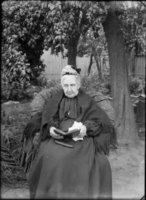 Outdoors backyard area, an unidentified elderly woman sitting under a pear tree with glasses, shawl and lace cap reading a book, with glasses case in her lap, probably Christchurch region