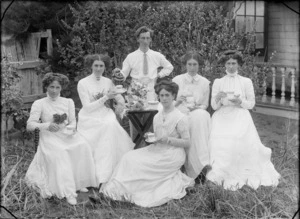 Group drinking tea in a garden, showing five unidentified women, holding teacups, served by an unidentified man pouring tea from a pot, and a small table laid with flowers, a plate of small cakes, a milk jug and sugar bowl, possibly Christchurch district