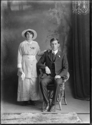 Studio portrait of an unidentified man and woman, showing the woman in a creased suit with a straw hat, and holding a pair of gloves, possibly Christchurch district