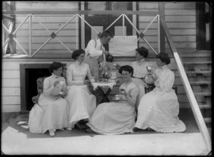 Tea party outdoors, showing five unidentified women eating small cakes and being served tea by an unidentified man, with a sign reading 'Golden Link Encampment, New Brighton, 1910' on balcony railing of wooden building behind