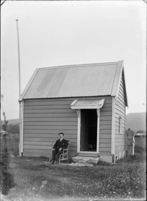 View of small hut with flag pole, with an unidentified man in three piece suit, imperial shirt collar and patterned tie sitting outside in a chair, probably Christchurch region