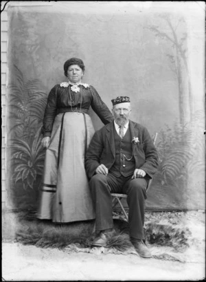 Outdoors portrait, an unidentified older couple in front of false backdrop, man with goatee beard and fez hat, wife with embroidered flower shaped collar over dark blouse, probably Christchurch region