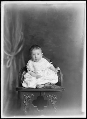 Studio portrait of an unidentified baby, sitting on a carved wooden chair, possibly Christchurch district