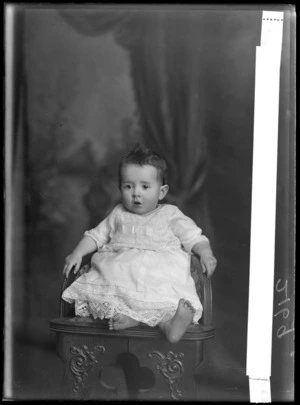 Studio portrait of unidentified baby in lace cotton top over crocheted under garment, sitting on wooden high chair, Christchurch