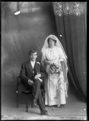 Studio unidentified wedding couple portrait, bride with very long veil holding flowers standing beside groom sitting, Christchurch