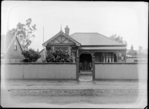 Street view of a single story wooden house with flag pole, single chimney, veranda and corrugated iron front fence and roof, probably Christchurch region
