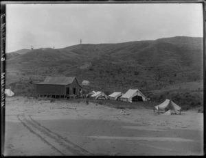 Mangakuri Beach with woolshed and tents, woman on beach with baby playing, hilly farmland beyond, Patangata, Hawke's Bay District