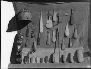 A display of Maori artifacts particularly patu, also tiki, axe heads, a walking stick, hooks, a pot, and a cloak