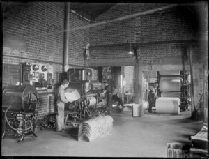 A group of unidentified men working with press machinery, with a man standing next to a paper roll machine, a man working on a press machine, and a man proof reading, possibly Hastings region