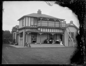 Members of an unidentified family sitting on the porch of a two storey wooden house, showing a baby carriage and a bicycle on the left side of the house and stairs on the right side of the house going up to the second floor, Hastings