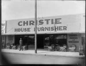 An unidentified man standing in front of the Christie House Furnisher store, Hastings, showing a variety of prams and strollers along the store front