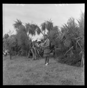 Featherston centennial celebrations, showing an unidentified man wearing traditional Maori costume, standing outdoors, with a group of unidentified girls standing behind a flax bush [during a ceremony?]