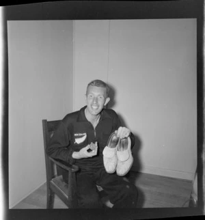 An unidentified man wearing a tracksuit with silver fern insignia on pocket, sitting in a chair, and holding [cricket?] shoes and a medal