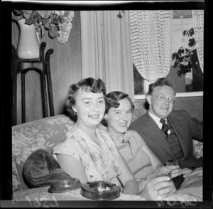 Unidentified people at an event for the Overseas Society, showing two women and a man sitting on a sofa, holding drinks, with ashtrays balancing on the armrest of chair in foreground