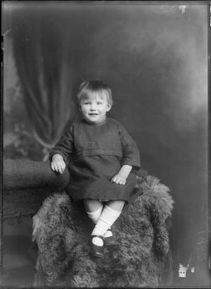 Studio portrait of unidentified young girl in a dark dress, white socks and leather shoes, sitting on a fur rug, Christchurch