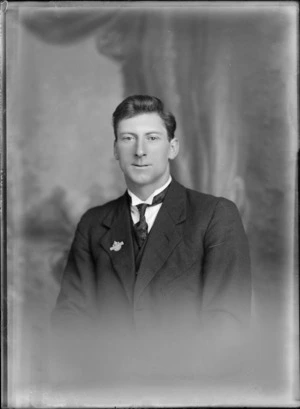 Studio upper torso portrait of an unidentified young man in a dark suit and a high wing stiff shirt collar with a dark patterned tie and lapel crown badge, Christchurch