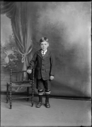Studio portrait of unidentified young boy in school uniform, with black and white striped tie and socks, Christchurch