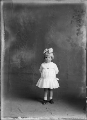 Studio portrait of unidentified young girl with a large hair bow and embroidered cotton dress with a waist bow, Christchurch