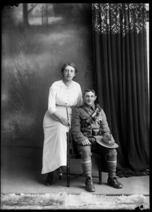 Studio portrait of unidentified couple, woman with glasses and large lace collar and striped skirt, sitting with her WWI soldier husband with bandolier shoulder and waist ammunition belts, stirrups and riding crop [Mounted Rifles?] and hat with badge, Christchurch