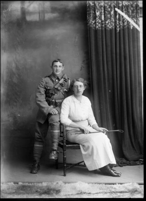Studio portrait of unidentified couple, woman with glasses, large lace collar and striped skirt, sitting with her WWI soldier husband with bandolier shoulder and waist ammunition belts, stirrups and riding crop [Mounted Rifles?], Christchurch