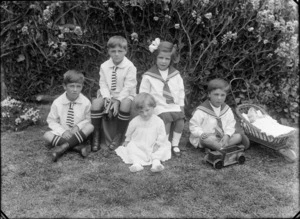 Outdoor portrait of unidentified children playing, two boys in school striped ties and socks, a boy and a girl in sailor tops and a toddler girl in lace dress. Toys include tin truck and train, a toy wooden pram with doll, probably Christchurch region