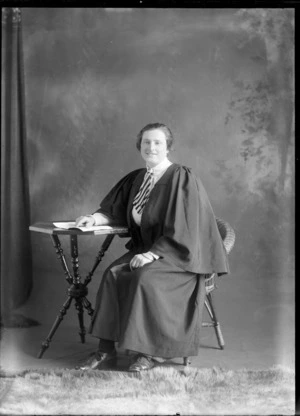 Studio portrait of unidentified young woman in a graduation gown with a striped necktie sitting at a table with a book, Christchurch