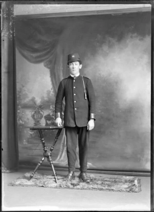 Studio portrait of unidentified young man in a [tram or train?] conductor's uniform with a single breasted dark jacket and pants, hat with '66' and a whistle hanging from his shoulder, Christchurch