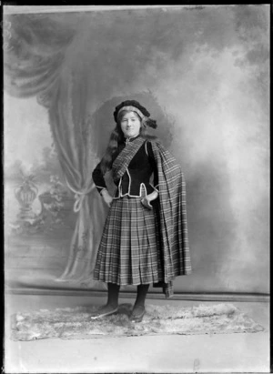 Studio portrait of an unidentified girl standing on a fur rug, dressed in a traditional Scottish costume, pleated tartan skirt, tartan scarf wrapped around her upper torso, draped and hanging off her left shoulder, wearing a velvet jacket and cap, possibly Christchurch district