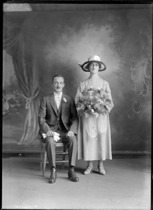 Studio portrait of an unidentified couple, showing the bride wearing a chiffon dress, with embroidery satin panels on the front of the dress, wearing a satin fur trim hat with floral adornments, holding a bouquet of flowers, showing the groom sitting on a wooden chair, dressed in a three piece suit, with a striped shirt, with a high collar and bow tie, with a posy in the button hole of his jacket, holding his gloves in his right hand, possibly Christchurch district