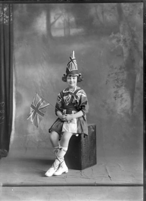Studio portrait of an unidentified girl in costume, showing the girl wearing a dress with hat made out of a Union Jack flag, with red, white, blue ribbons over her long white socks, and holding a flag, sitting on a wooden crate, possibly Christchurch district