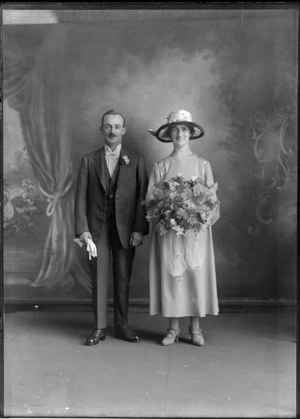 Studio wedding portrait of an unidentified couple, showing the bride wearing a chiffon dress, with embroidery satin panels on the front of the dress, wearing a satin fur trimmed hat with floral adornments, holding a bouquet of flowers, showing the groom dressed in a three piece suit, with a striped shirt, with a high collar and bow tie, with a posy in the button hole of the jacket, holding his gloves in his right hand, possibly Christchurch district