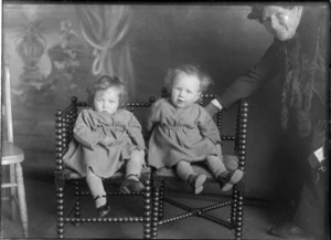 Studio portrait of two unidentified [twin?] girls sitting on wooden chairs, dressed in heavy cotton dresses, woollen socks, black button shoes, showing a partially obscured unidentified woman on the rightside of the children, holding one of the girls up, possibly Christchurch district