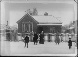 Outdoor portrait of two unidentified women with children in front of a fence, in front of a brick house, on a snow covered unidentified street, showing a girl, a boy holding lumps of snow in their right hand, showing a younger boy standing alongside a partially obscured [girl?] on the rightside, probably Christchurch district