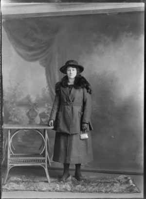 Studio portrait of an unidentified woman wearing a two piece woollen skirt with coat, with a fur stole draped over her shoulders, holding a coin purse in her left hand, standing on a fur rug next to a cane table with he right hand resting on the table top, possibly Christchurch district