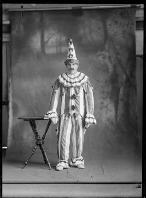 Studio portrait of an unidentified man dressed in dress up costume, wearing a striped clown suit with pompoms on the top, three pompoms down the middle of his pointed hat, possibly Christchurch district