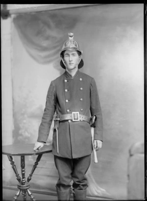 Studio portrait of an unidentified man wearing a fireman's suit with helmet, tool belt with spike tool, axe, possibly Christchurch district