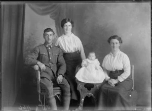 Studio portrait of members of an unidentified family, showing the man wearing his army uniform sitting on a cane seat, with a woman standing next to him, with a infant child dressed in a smocked gown sitting on a wooden high chair next to a woman sitting on a wooden seat, possibly Christchurch district