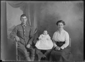 Studio portrait of members of an unidentified family, showing the man dressed in his army uniform sitting on a cane seat, with the infant child dressed in a smocked gown sitting on a wooden high chair, and the woman sitting next to the child, possibly Christchurch district