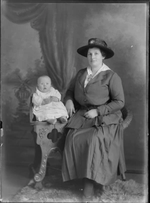 Studio portrait of an unidentified woman with a infant child, showing the child wearing a white cotton lace smocked gown, and the woman dressed in a two piece skirt suit, with a white cotton blouse, a floral posy pinned on the front of her blouse and a silk hat, possibly Christchurch district