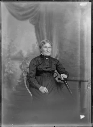 Studio upper torso portrait of an unidentified woman sitting on a cane seat wearing a dark dress with a brooch on the high neck collar, showing the woman resting her left arm on a wooden table, possibly Christchurch district