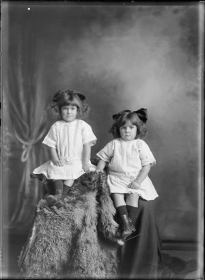Studio portrait of two unidentified [twin?] sisters, wearing cotton dresses with belts, with large ribbons in their hair, showing the girls sitting on a fur rug covered high stools, possibly Christchurch district