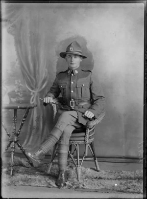 Studio portrait of an unidentified man dressed in his army uniform holding a cane, sitting on a cane seat next to a wooden table on a fur rug, possibly Christchurch district
