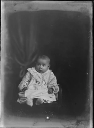 Studio portrait of an unidentified infant child dressed in a cotton gown, with a large lace collar, a matinee jacket with ribbons, with white tights, showing the infant sitting on a wooden high chair, possibly Christchurch district