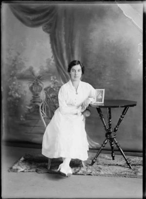 Studio portrait of an unidentified woman sitting on a cane chair on a fur rug, showing the woman dressed in a [nurses uniform?], with a brooch attached to the neckline, showing the woman leaning on the table with a framed photograph of an unidentified woman, possibly Christchurch district