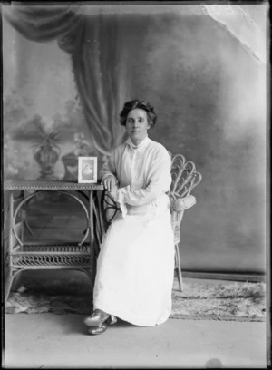 Studio portrait of an unidentified woman wearing a [nurses uniform?], with a cream coloured jacket with frilly sleeves, showing the woman sitting on a cane chair next to a cane table with a photograph in a frame of an unidentified woman, possibly Christchurch district