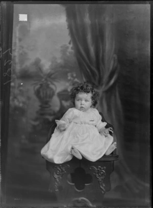Studio unidentified family portrait of a baby in a christening gown with a greenstone bar brooch sitting on a wooden highchair, Christchurch