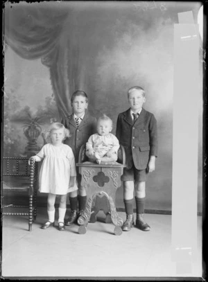 Studio unidentified family portrait of four children, with two older boys in jackets, pants and school tie, and a young girl standing with a baby on a highchair, Christchurch
