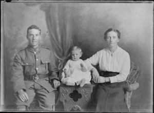 Studio unidentified family portrait of a young World War I soldier with fern collar badges and his wife in a lace cotton shirt, sitting beside their baby on a wooden highchair, Christchurch