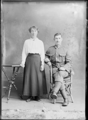 Studio unidentified family portrait of a young World War I soldier with moustache and fern collar badges, putties and hobnail boots, sitting holding a swagger stick next to his wife standing, Christchurch